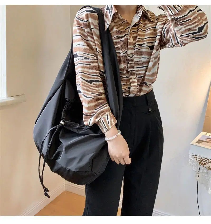 Porté à l'épaule sur une chemise imprimée, le grand sac noir se cale sous le bras, bien rempli
