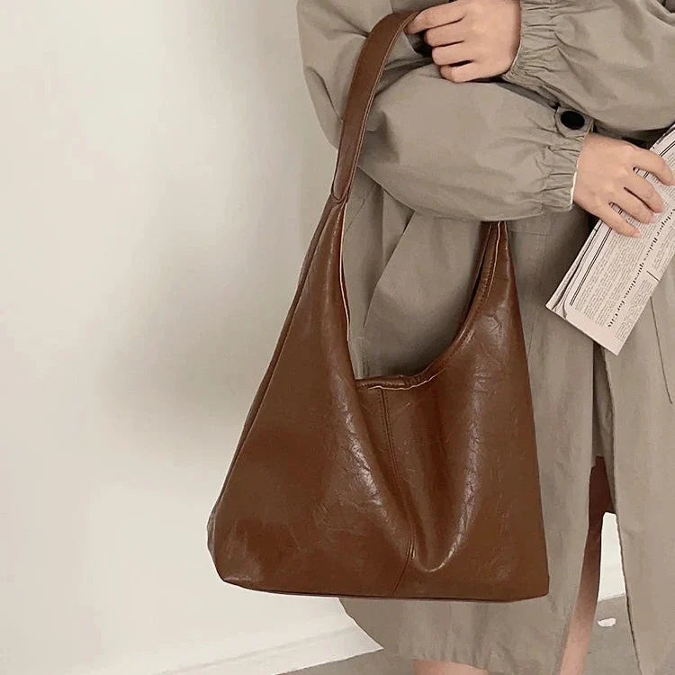 Anse glissée sur l'avant-bras, le sac cognac se détache d'un manteau sable, journal à la main