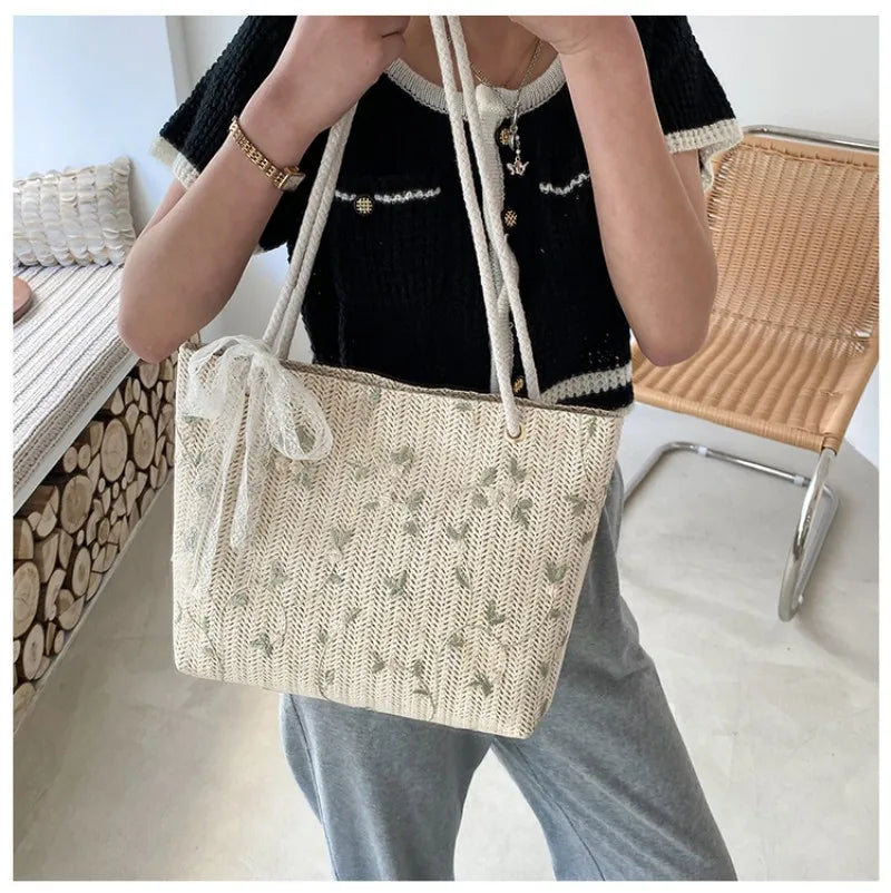 Porté à l'épaule sur un gilet noir, le cabas tressé beige laisse pendre son nœud de dentelle