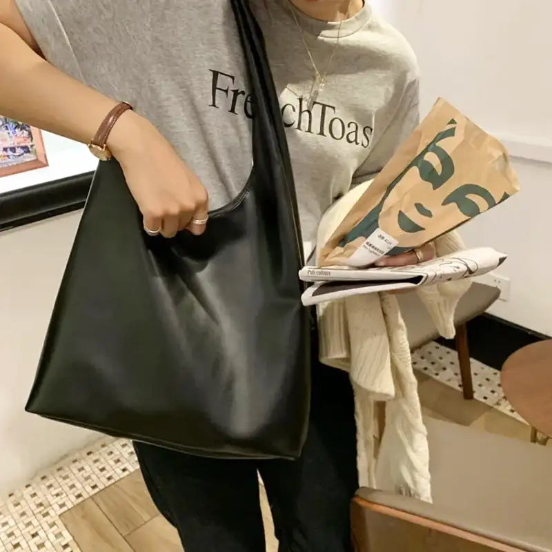Sac noir tenu à la main devant un tee-shirt gris, avec un journal et un sac en papier kraft