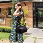 Porté en croisé sur une robe fleurie verte, le sac noir repose sur la hanche, en extérieur