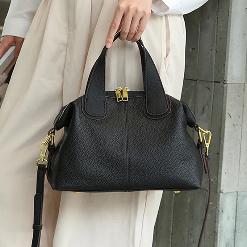Porté à la main, le sac noir grainé se détache sur une tenue écrue, sangle réglable bien visible