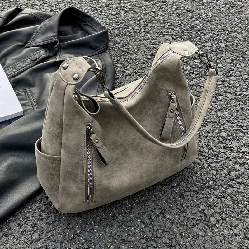 Posé sur le gravier près d'un blouson noir, le sac kaki grisé montre ses deux zips obliques