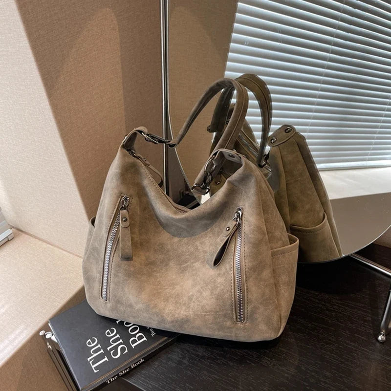 Brun taupe et souple, le sac repose sur des livres devant un store, tirettes métal apparentes