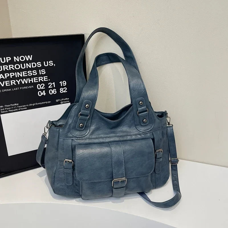 Posé sur un plateau blanc devant une affiche, le sac bleu ardoise laisse voir sa bandoulière
