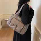 Épaulé le long d'un manteau noir, le sac gris taupe montre son rabat bouclé et son anse plate
