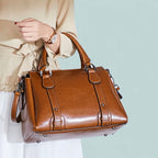 Tenu à la main devant un fond bleu pâle, le sac cognac accompagne une blouse beige