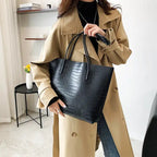 Le cabas noir croco tenu par les anses devant un trench camel, ouverture large et sans fermeture