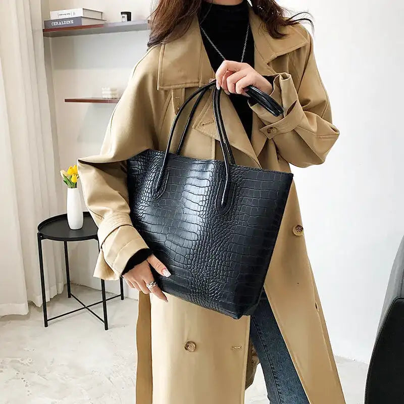 Le cabas noir croco tenu par les anses devant un trench camel, ouverture large et sans fermeture