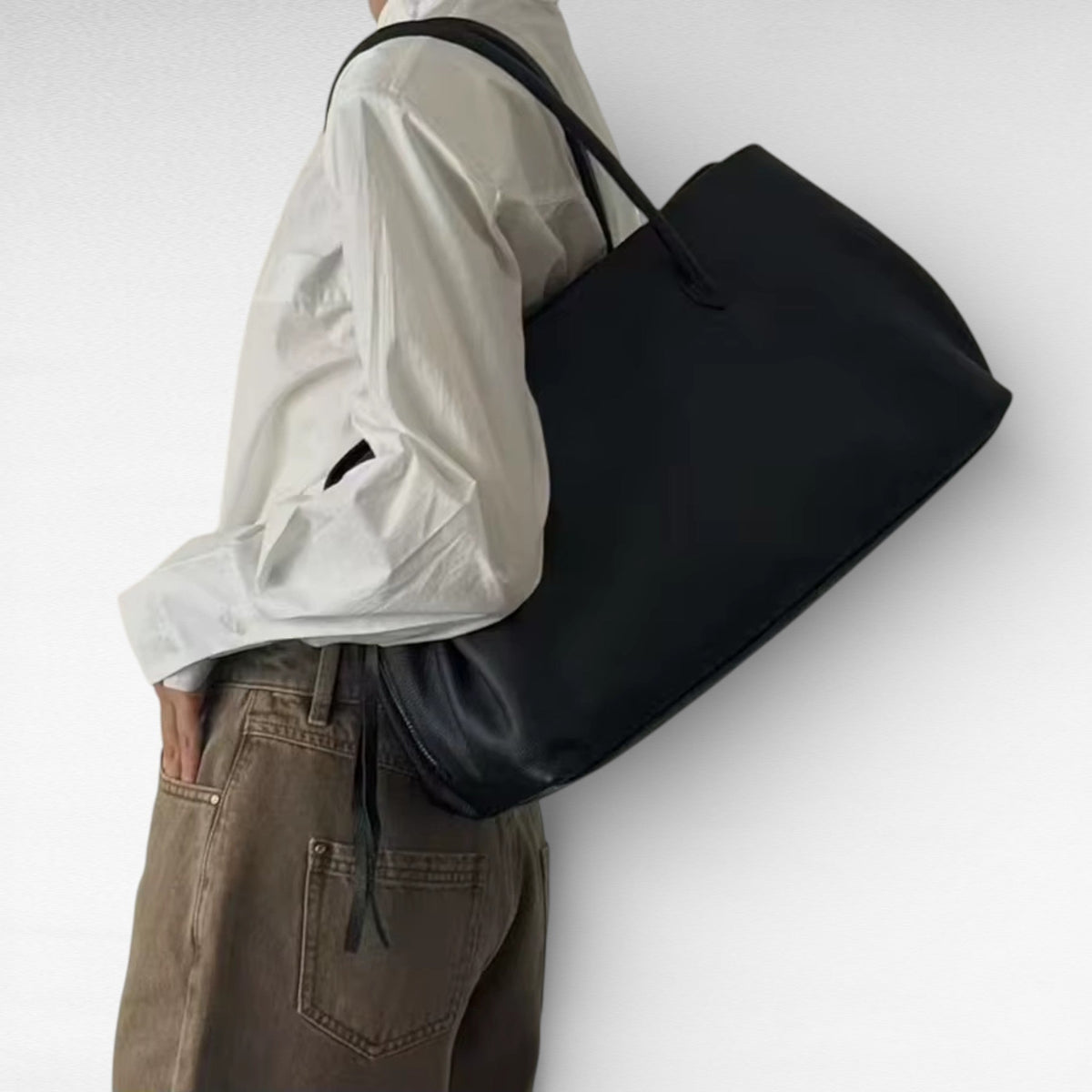 Porté à l'épaule, le cabas noir repose contre une chemise blanche et un pantalon marron