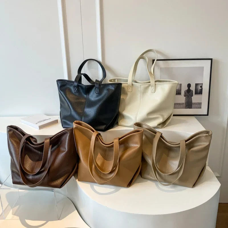 Cinq coloris du même cabas réunis sur un présentoir blanc : noir, crème, brun, caramel, taupe