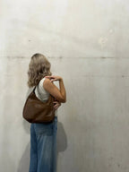 Adossée à un mur de béton, une jeune femme porte le sac marron à l'épaule sur un jean clair