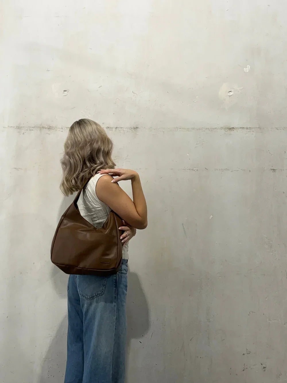 Adossée à un mur de béton, une jeune femme porte le sac marron à l'épaule sur un jean clair