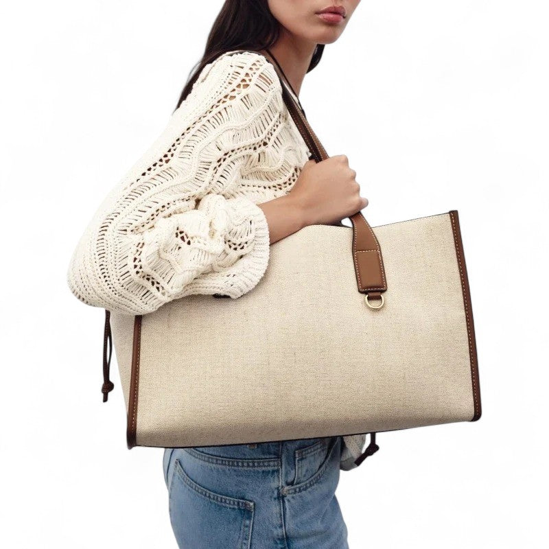 Sous le bras, sur un pull crocheté écru, le grand cabas en toile beige montre son flanc et sa bride