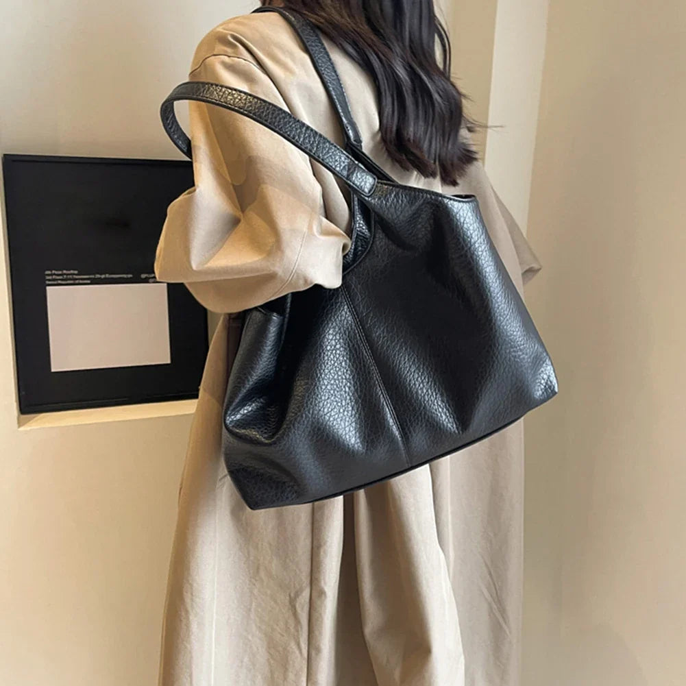 Porté à l'épaule sur un manteau beige, le cabas noir se creuse en plis souples le long du buste