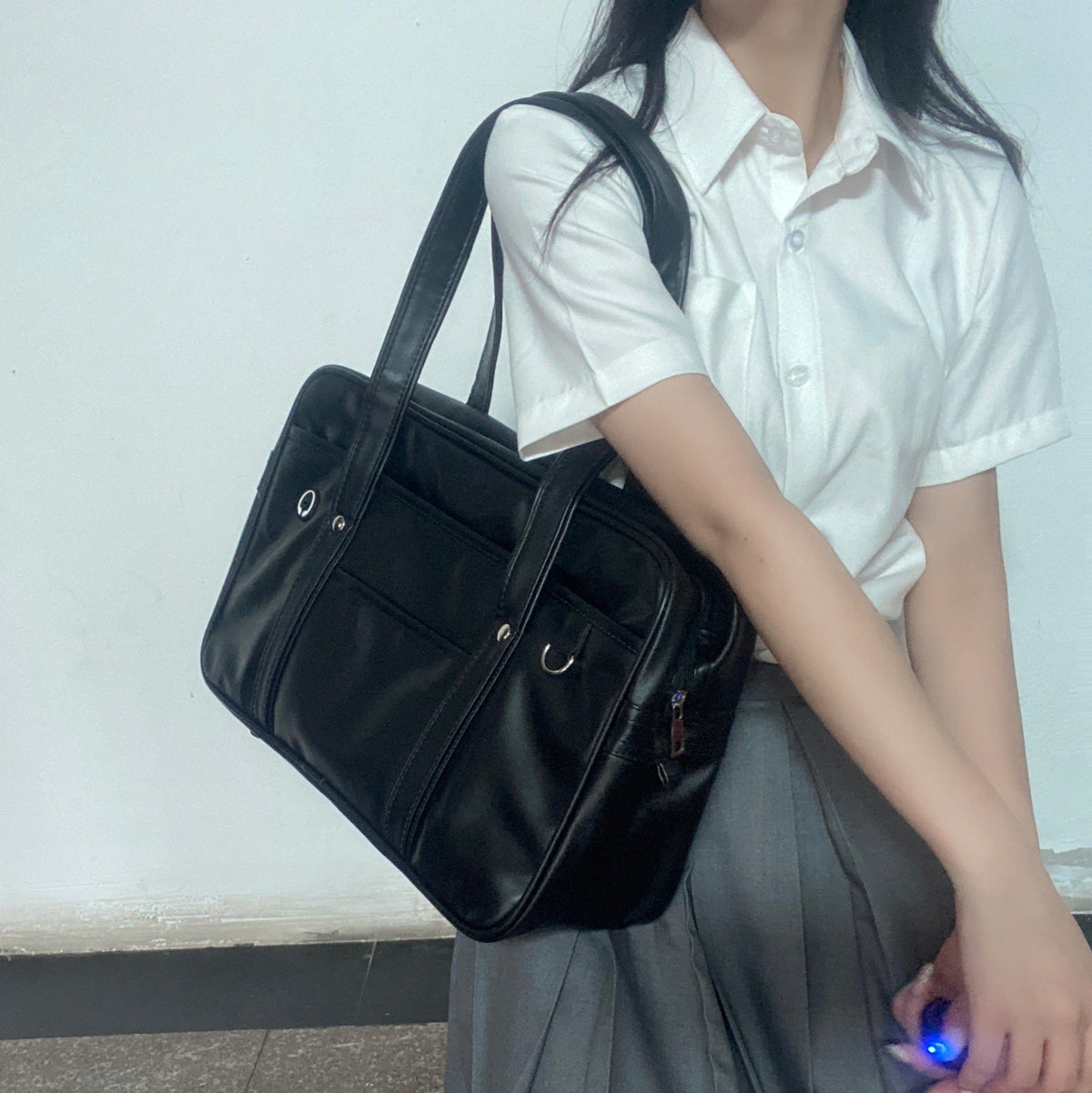 En extérieur, le sac noir passe sur l'épaule d'une lycéenne en chemisier blanc et jupe grise
