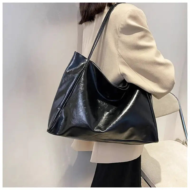 À l'épaule sur un manteau beige, le cabas noir s'aplatit contre la hanche, anses regroupées en main