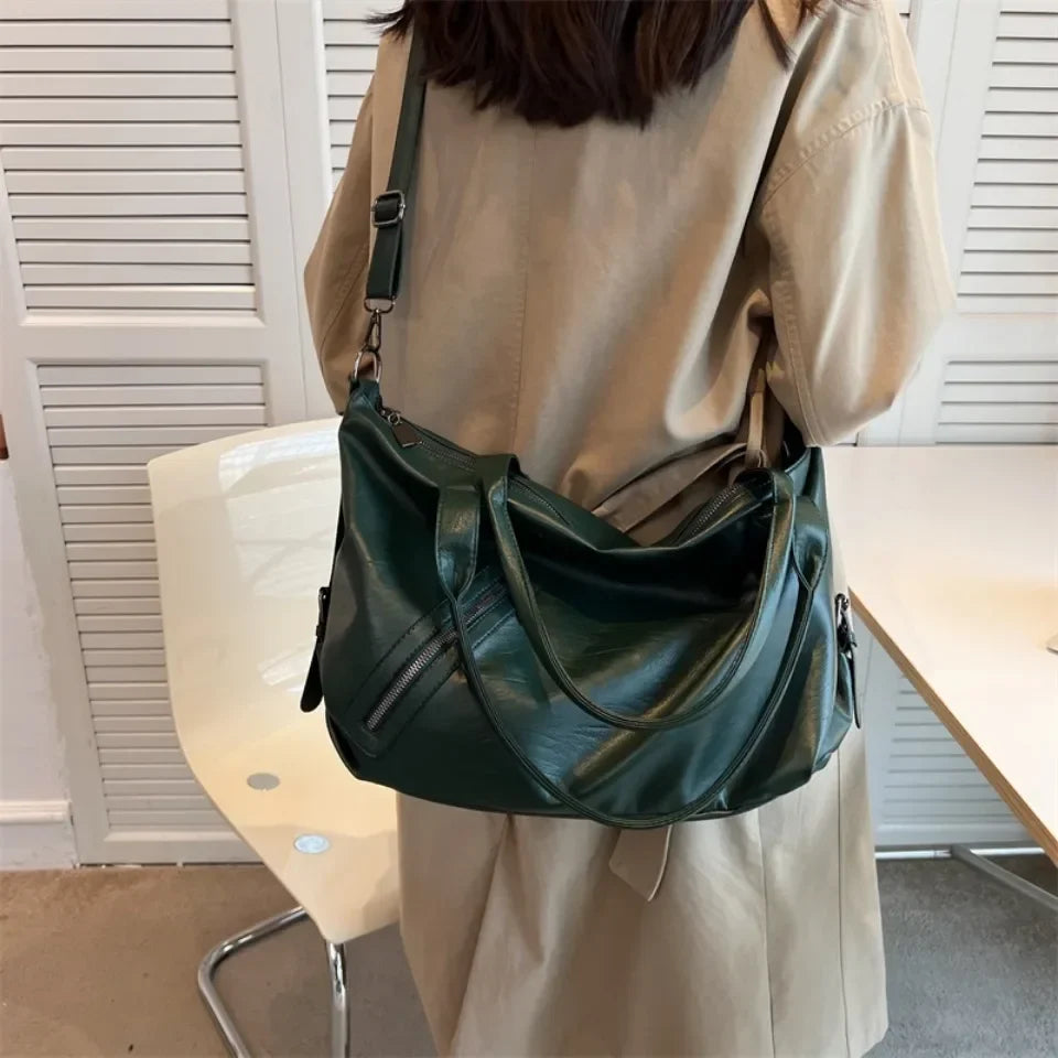 Bandoulière passée en travers du dos, le sac vert foncé repose sur la hanche d'une silhouette en beige