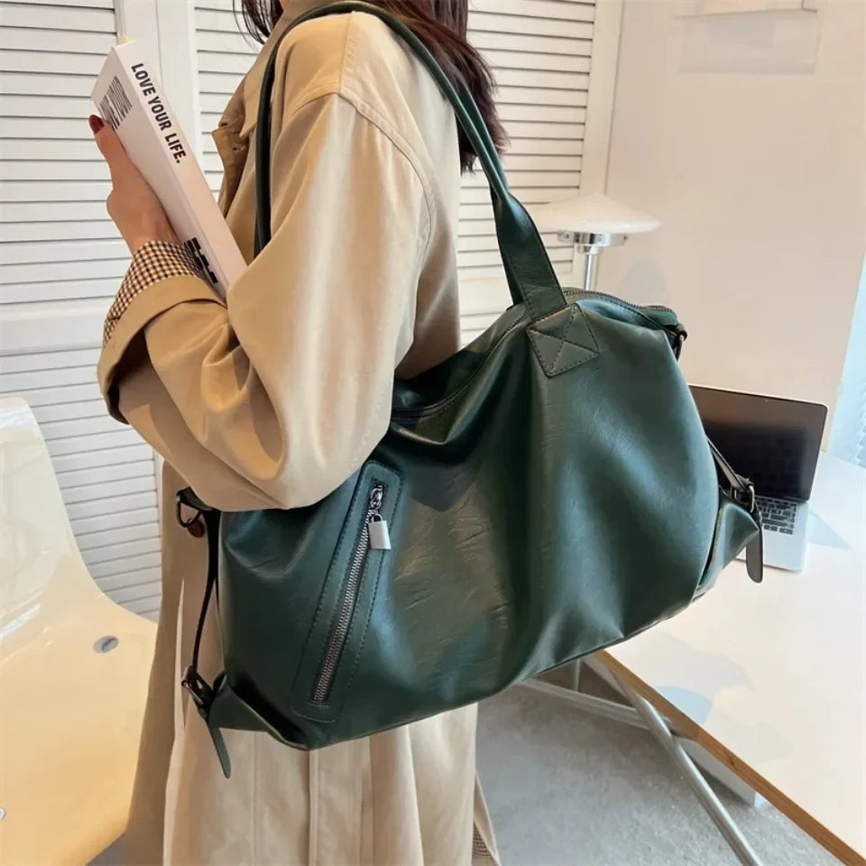 Épaule chargée du sac vert foncé, tenue beige et magazine à la main, dans un intérieur très clair