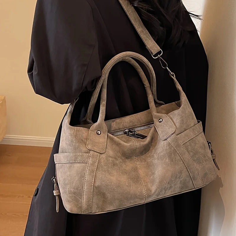 Porté à la main contre un manteau sombre, le sac taupe s'affaisse en accordéon, zip entrouvert