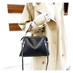 Silhouette en trench beige tenant la besace noire, bandoulière fine retombant librement