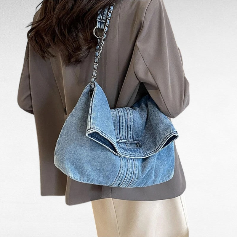 De dos, la besace en jean repose sur l'épaule d'une veste taupe portée avec un pantalon crème