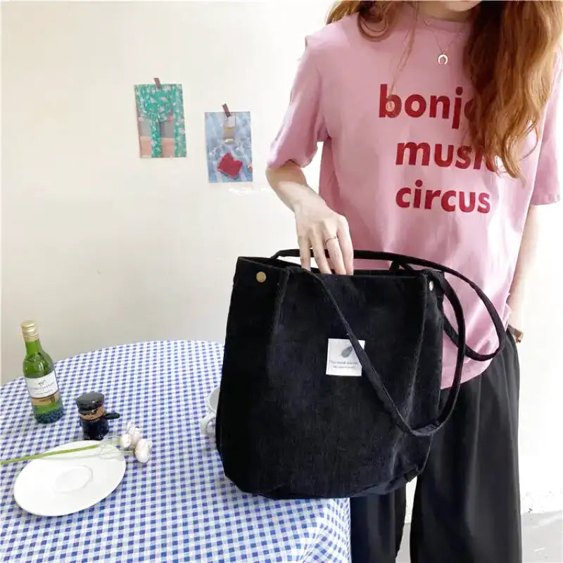 Main saisissant les anses du tote bag noir côtelé, devant une table nappée de vichy bleu