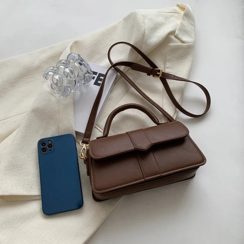 Format compact du sac brun, posé près d'un téléphone bleu sur une veste beige froissée