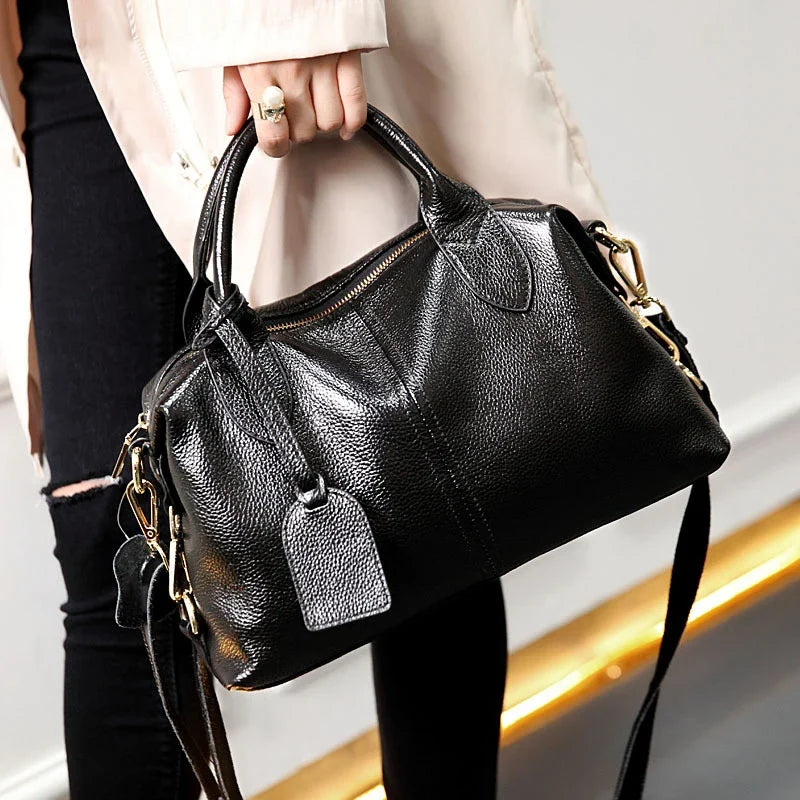 Tenu à la main devant un jean noir, le sac grainé laisse pendre sa bandoulière et son étiquette