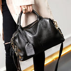 Tenu à la main devant un jean noir, le sac grainé laisse pendre sa bandoulière et son étiquette