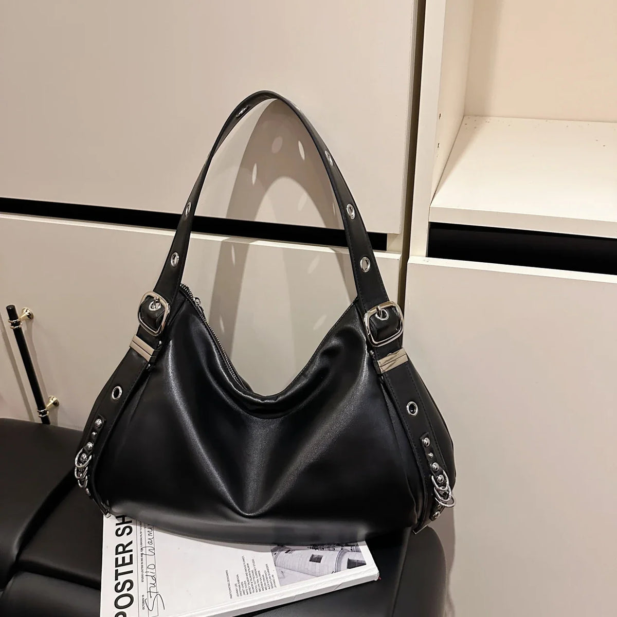 Sur un meuble clair près d'un magazine, le sac noir laisse voir ses œillets et sa boucle