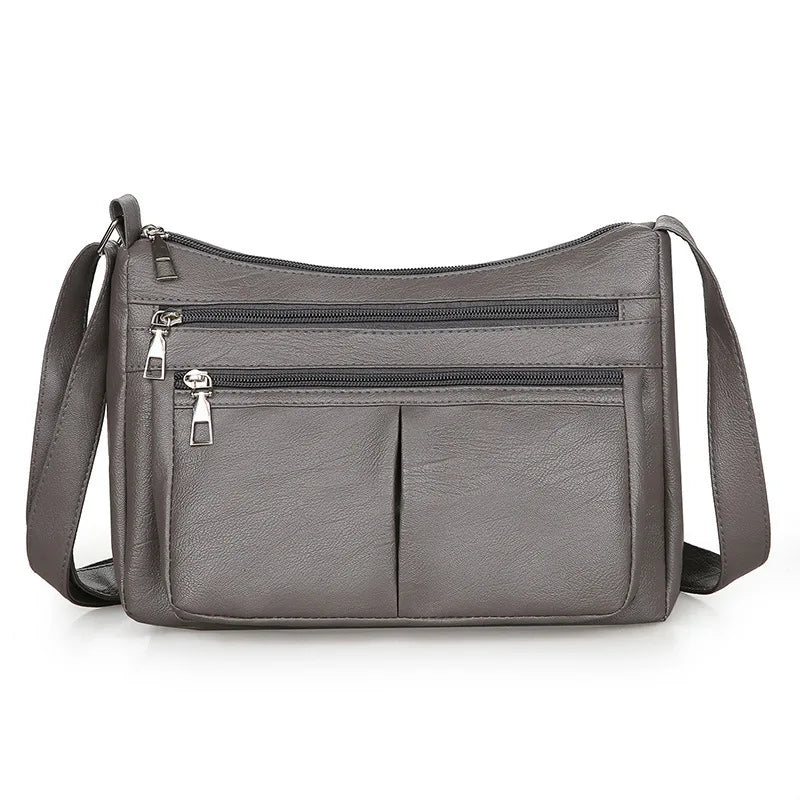 Gris souris mat, le sac bandoulière montre ses deux glissières et sa poche soufflée centrale