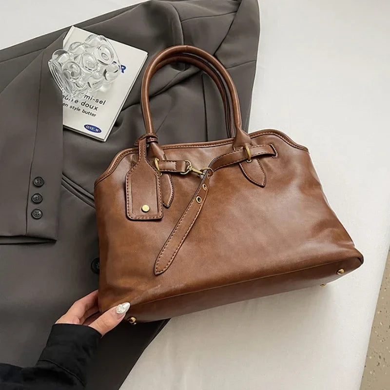 Cuir végan cognac patiné, le cabas posé de trois quarts sur une veste grise, base cloutée visible