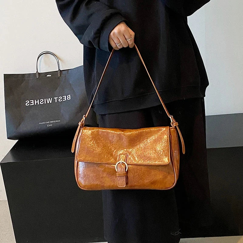 Tenue à la main devant un sac en papier, la besace cognac brille de son grain lustré, rabat fermé