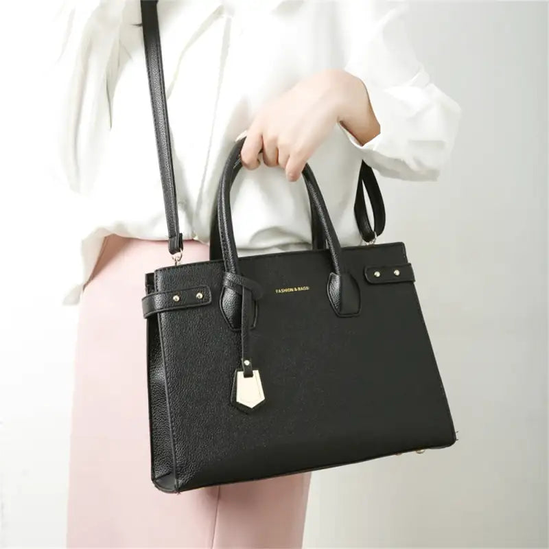 Main tenant le sac noir par ses anses rigides, sur une chemise blanche et un pantalon rose