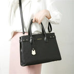 Main tenant le sac noir par ses anses rigides, sur une chemise blanche et un pantalon rose