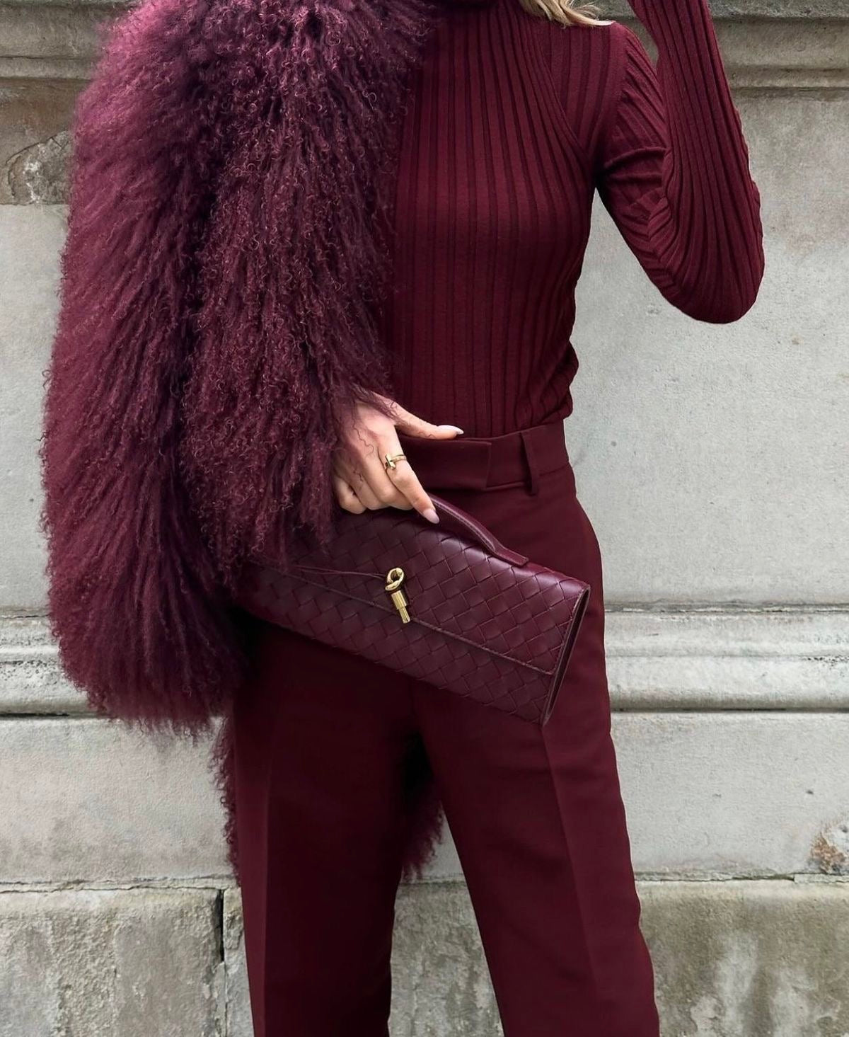 Tenue à la main devant une veste duveteuse prune, la pochette bordeaux tressée capte la lumière