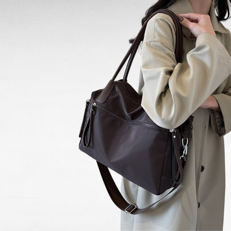Épaule chargée du sac brun foncé, dont la bandoulière pend le long d'un manteau beige