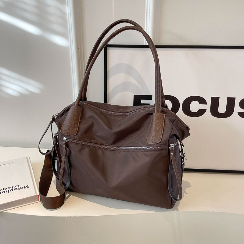 Grand sac brun posé sur un socle blanc, vu de trois quarts, poches zippées latérales visibles