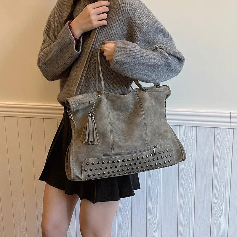 Tenu au creux du bras sur un pull gris, le cabas taupe clouté s'ouvre par une glissière