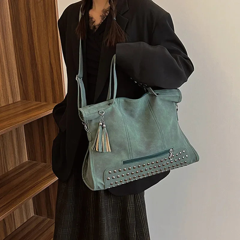 Vert de gris clouté, le grand sac se porte en bandoulière par-dessus une veste noire