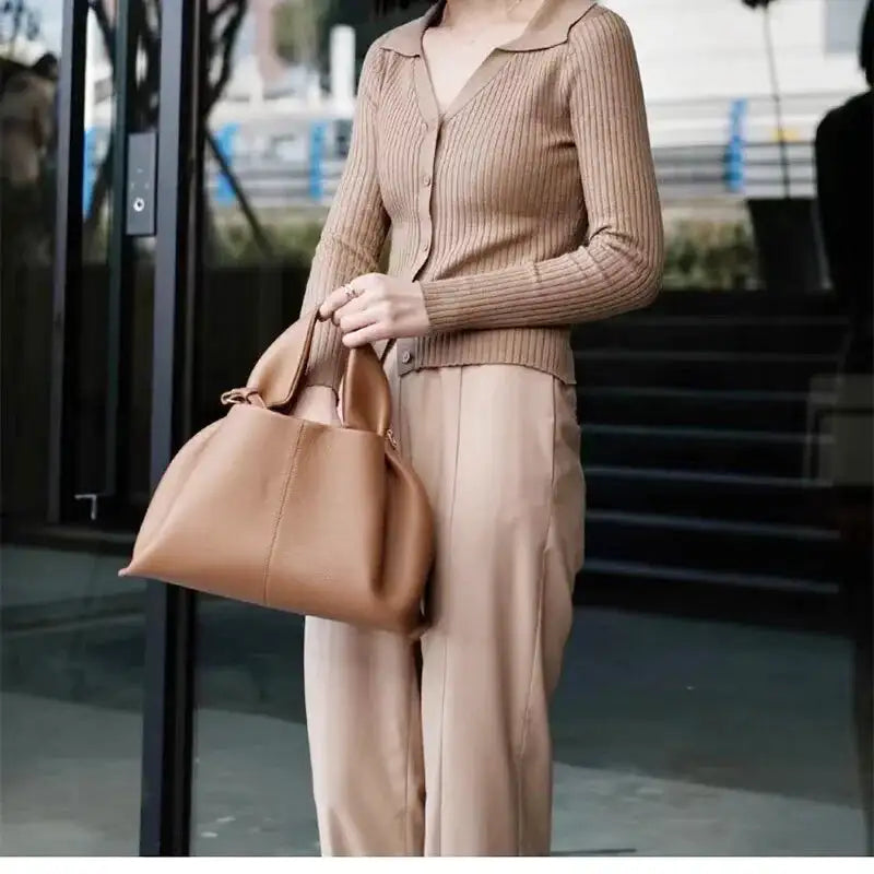 Femme en cardigan beige tenant le sac camel à la main devant une façade vitrée, plis marqués