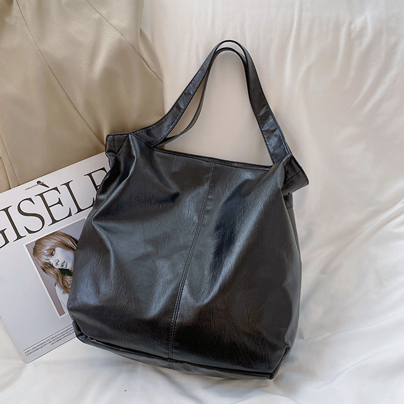 Noir lustré et matière lisse, le sac fourre-tout repose sur un drap blanc près d'un magazine