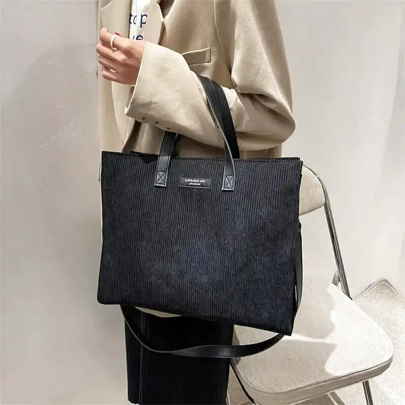 Épaule d'un manteau clair supportant le cabas noir côtelé, sangle retombant le long du corps