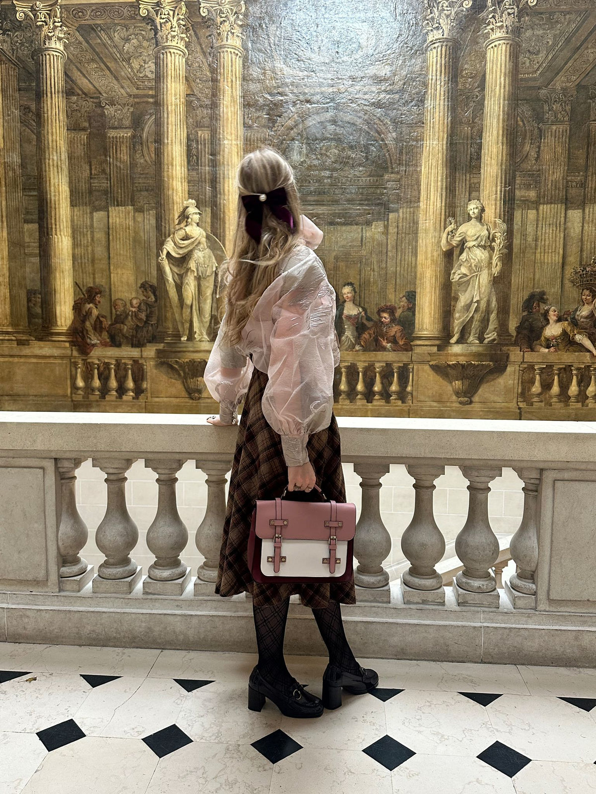 De dos dans une salle de musée, une femme tient le cartable rose et bordeaux à la main