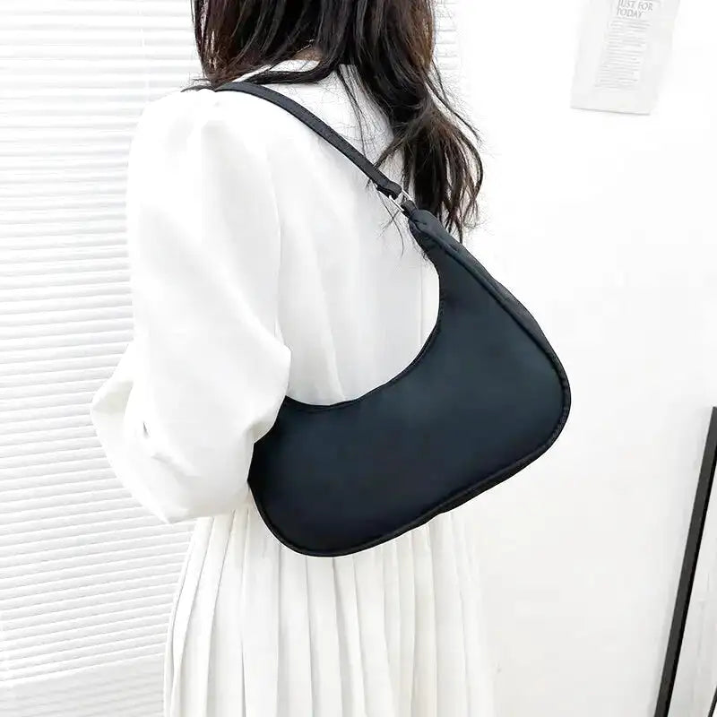 De dos, le sac noir repose sur l'épaule d'une femme en chemise blanche et jupe plissée