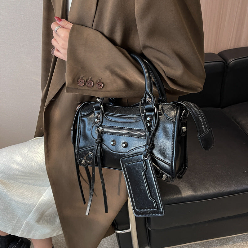 Glissé sous le bras d'une veste marron, le sac noir lustré montre son zip et ses œillets