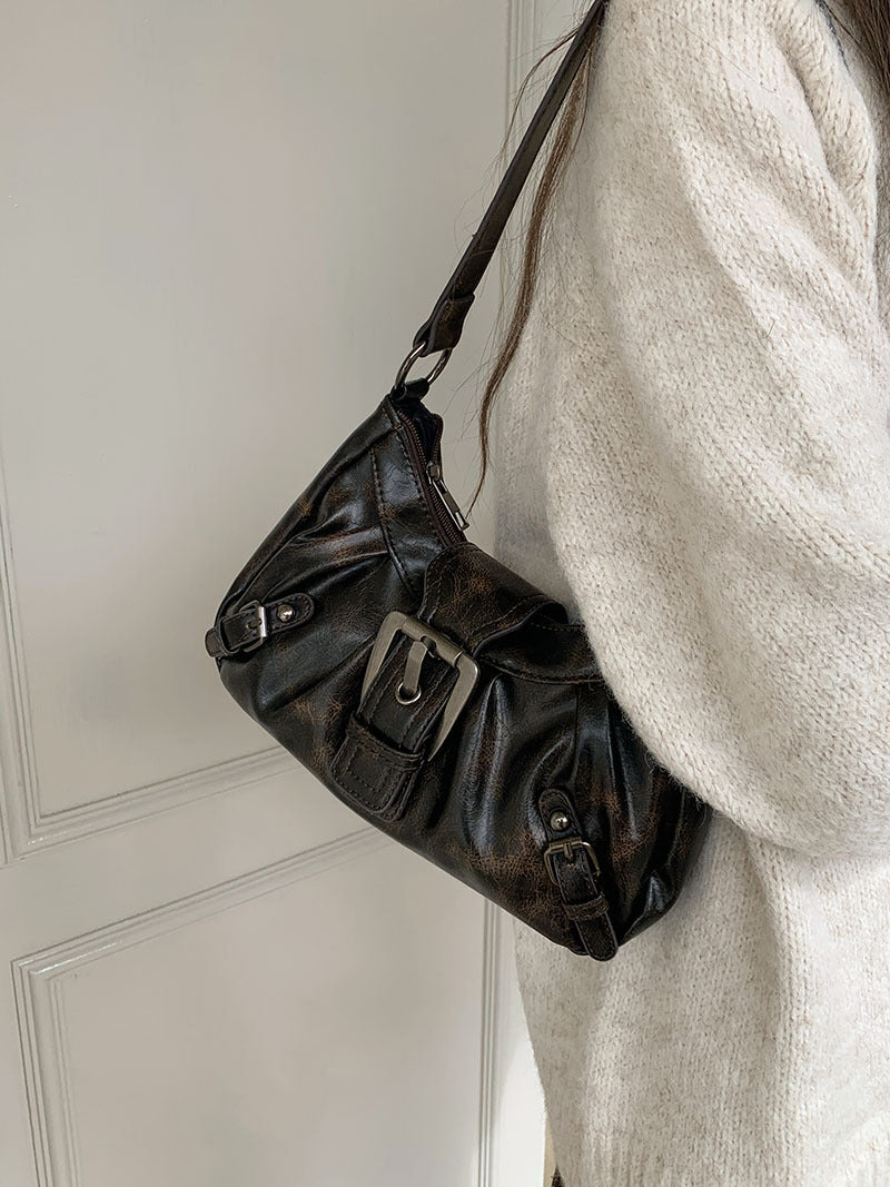 De biais contre un mur clair, le sac brun posé sur l'épaule laisse voir sa boucle latérale