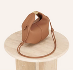 Posé sur un guéridon de pierre claire, le sac camel laisse tomber sa longue sangle nouée