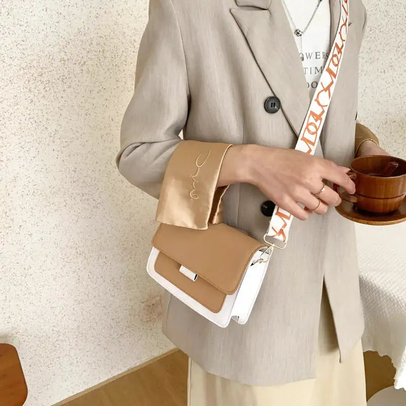Main posée sur le sac camel et blanc porté en bandoulière, blazer beige et mug en bois en arrière-plan